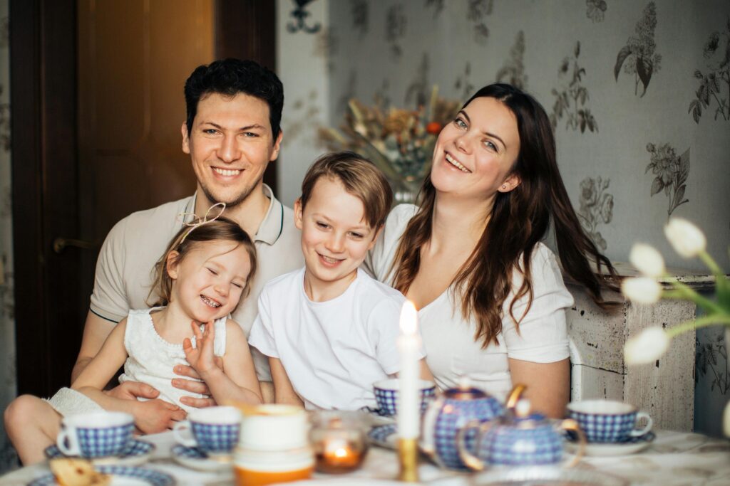 Happy family laughing while children sitting on laps at table set with tea and flowers near burning candle